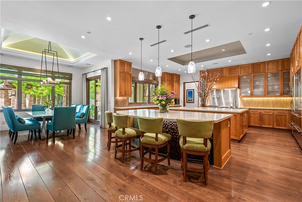 This spacious, high-end kitchen features warm wood cabinetry, a large central island with bar seating, and a dedicated dining area to the left. The room is illuminated by elegant pendant lighting and recessed lights, highlighting the polished hardwood floors and clean, sophisticated design. The perspective is a wide-angle shot from the kitchen floor, showcasing the seamless flow between the cooking and dining spaces.