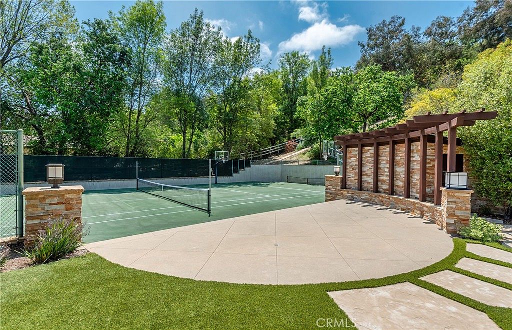 This expansive outdoor space features a private tennis court set against a backdrop of lush, mature trees and a hillside. A stone-accented pergola provides a shaded seating area on a concrete patio, seamlessly transitioning into a well-manicured lawn with stone pavers. The scene captures a serene and luxurious recreational area perfect for outdoor activities and entertaining.