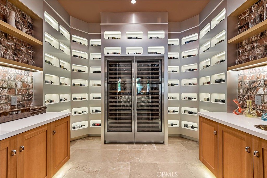 This sophisticated wine cellar features a symmetrical design with a central stainless steel dual-door wine refrigerator flanked by custom built-in shelving for individual bottle storage. The space is accented by warm under-cabinet lighting, rustic brick wall sections, and light-toned stone countertops, creating a luxurious and inviting atmosphere. The perspective is a centered, eye-level shot that emphasizes the organized, high-end storage capabilities of the room.
