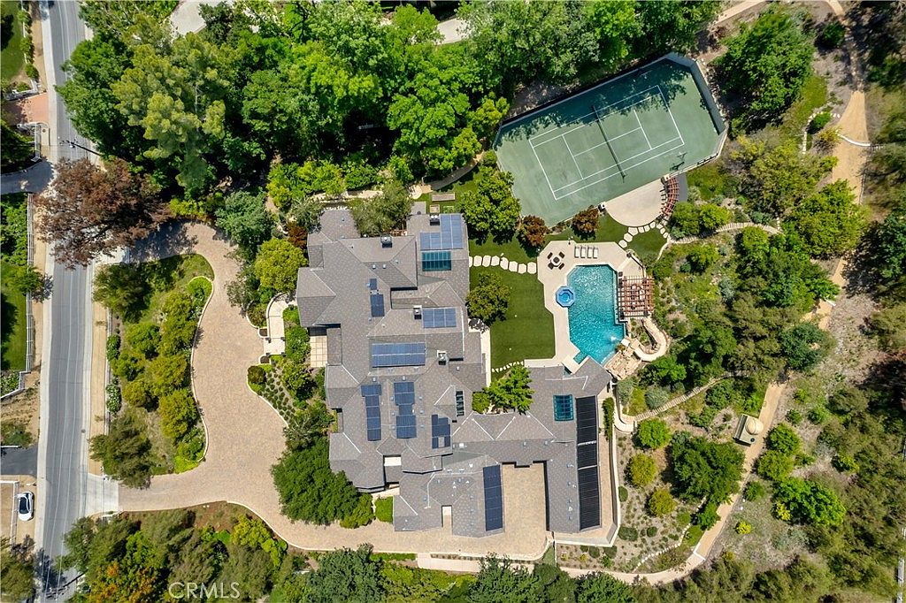 This high-angle aerial view captures a sprawling luxury estate nestled within a lush, wooded landscape. The property features a large multi-gabled residence with solar panels, a private tennis court, and a resort-style swimming pool with an integrated spa and rock waterfall. A winding paved driveway leads to the home, which is surrounded by mature trees and manicured landscaping, conveying an atmosphere of privacy and exclusivity.