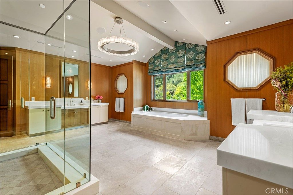 This luxurious primary bathroom features warm wood-paneled walls, a spacious walk-in glass shower, and a deep soaking tub positioned beneath a large window with patterned Roman shades. The room is anchored by a central crystal chandelier and light-colored stone flooring, creating an elegant and serene atmosphere. The perspective captures the expansive layout, highlighting the dual vanity areas and the sophisticated, high-end design.