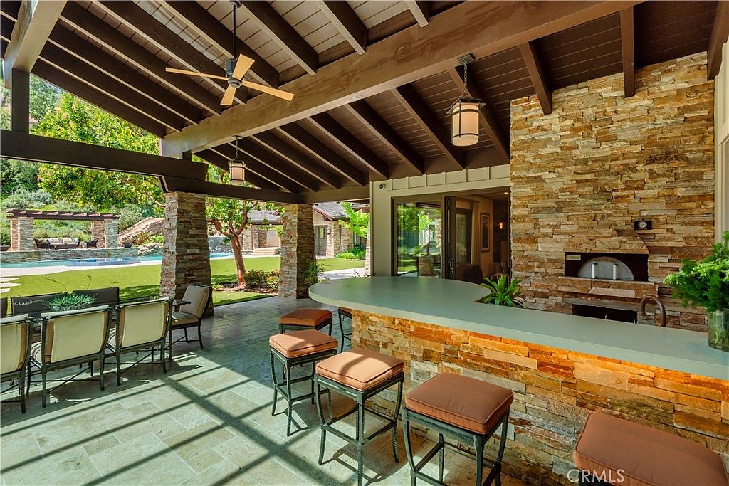 This expansive covered patio features a rustic stone-clad bar with a smooth countertop, perfect for outdoor entertaining. The space is equipped with a built-in pizza oven, ceiling fans, and comfortable bar seating, all overlooking a lush backyard with a swimming pool. The warm wood-beamed ceiling and stone pillars create a sophisticated, inviting atmosphere that seamlessly blends indoor and outdoor living.