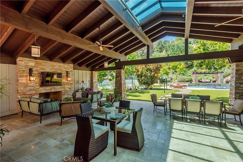 This expansive, covered outdoor living space features a rustic stone fireplace, a dining area, and comfortable lounge seating, all under a vaulted wood-beam ceiling with a skylight. The open-air design seamlessly transitions to a lush backyard with a swimming pool and manicured landscaping, creating an ideal environment for luxury entertaining. The perspective is from the center of the patio, looking outward toward the sun-drenched garden.