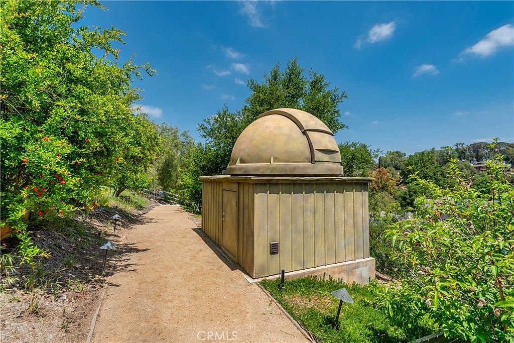 This unique outdoor space features a custom-built backyard observatory with a classic dome roof, set amidst a lush, landscaped garden. A gravel pathway leads directly to the structure, which is surrounded by mature trees and vibrant greenery under a bright blue sky. The setting offers a serene and private atmosphere, highlighting a rare and distinctive architectural amenity for stargazing enthusiasts.