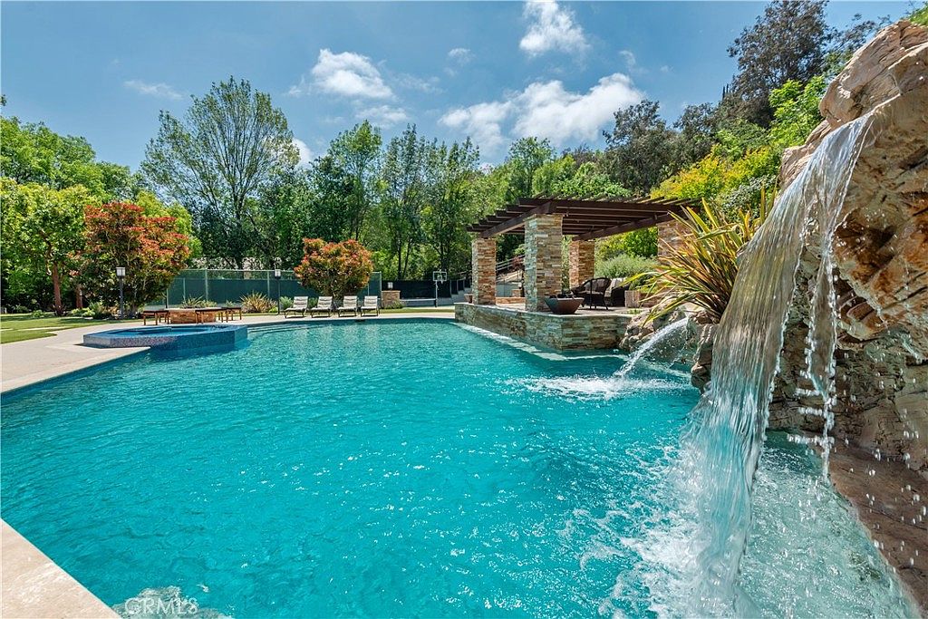 This luxurious backyard features a large, sparkling blue swimming pool with an integrated rock waterfall feature and a raised spa. A stone-pillared pergola provides a shaded lounge area, while the surrounding lush greenery and mature trees create a private, resort-like atmosphere. The perspective is captured from the poolside, emphasizing the expansive water feature and the serene outdoor living space.