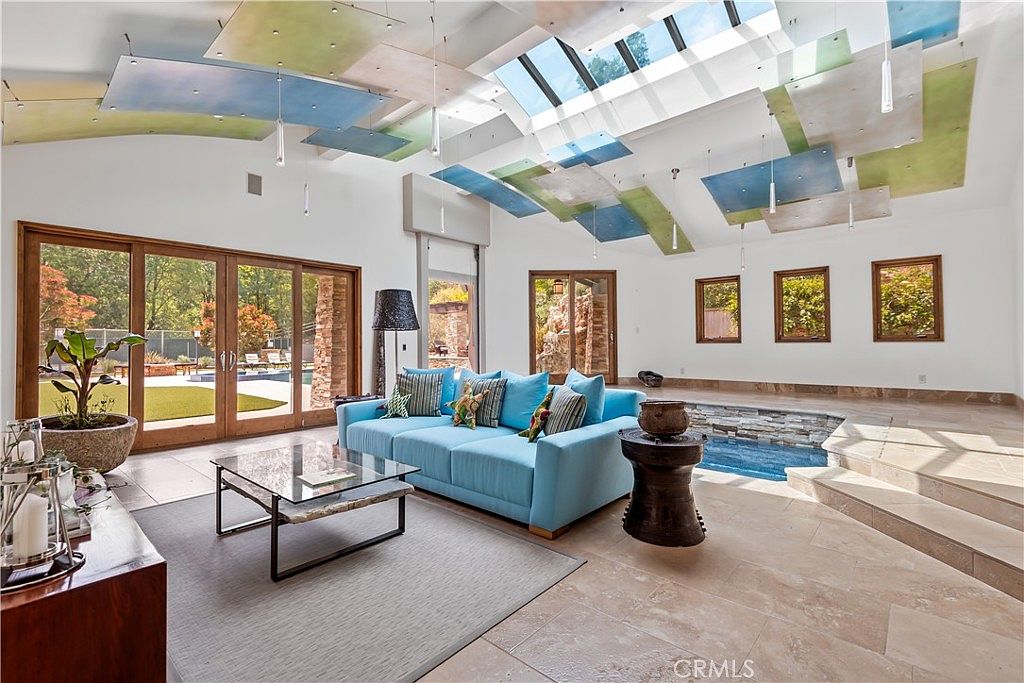 This expansive, high-ceilinged living room features a striking architectural design with suspended, multi-colored ceiling panels and a large skylight that floods the space with natural light. The room is anchored by a vibrant blue sofa and a sunken indoor water feature, creating a unique, resort-like atmosphere. Large glass doors provide a seamless transition to the outdoor pool area, emphasizing a luxurious, open-concept lifestyle.