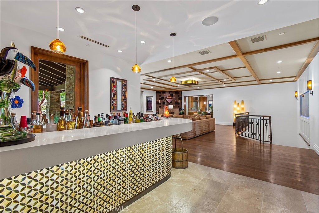 This sophisticated interior space features a prominent, curved wet bar with a decorative geometric tile front and a sleek white countertop, serving as the focal point of the room. Beyond the bar, the open-concept layout flows into a spacious living area with warm wood flooring, a coffered ceiling, and a cozy seating arrangement. The perspective captures the transition from the bar area into the main living space, highlighting the home's luxurious and inviting atmosphere.