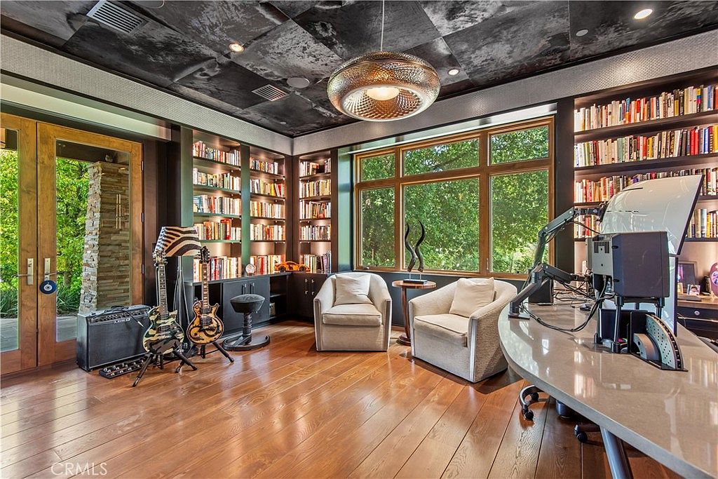 This sophisticated home office features rich hardwood flooring, extensive built-in bookshelves, and a unique textured ceiling with a large, modern pendant light. The room is furnished with two comfortable armchairs, a sleek desk setup, and musical instruments, creating a creative and professional atmosphere. Large windows provide ample natural light and a view of the lush greenery outside, enhancing the room's inviting and productive ambiance.