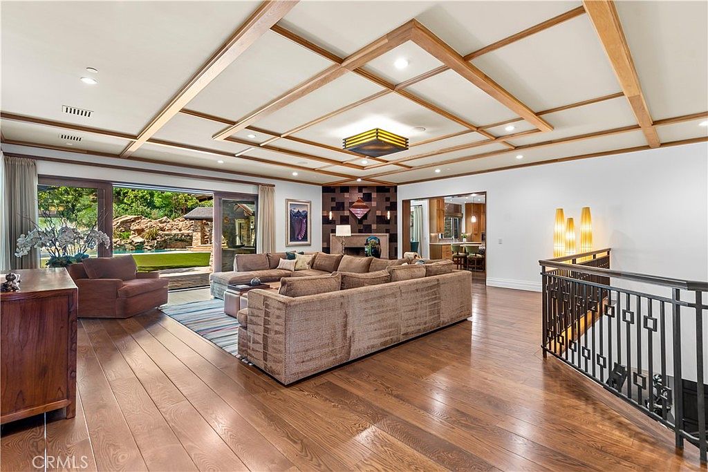 This spacious, open-concept living room features a coffered wood-beam ceiling and rich hardwood flooring, creating a warm and sophisticated atmosphere. A large, plush sectional sofa serves as the centerpiece, facing a unique geometric-tiled fireplace wall, while floor-to-ceiling sliding glass doors provide a seamless transition to the outdoor pool area. The perspective captures the expansive layout, highlighting the elegant architectural details and the inviting, airy flow of the home.