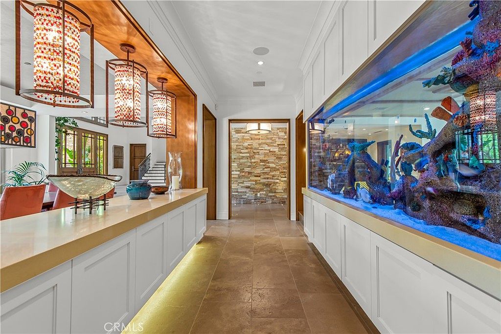 This elegant hallway features a striking, floor-to-ceiling built-in saltwater aquarium that serves as a dramatic focal point. The space is illuminated by three ornate, cylindrical pendant lights hanging from a warm wood-paneled ceiling, while the floor is finished in neutral stone tiles. A clean, white cabinetry base runs along the wall beneath the aquarium, leading the eye toward a stone-accented entryway in the background.