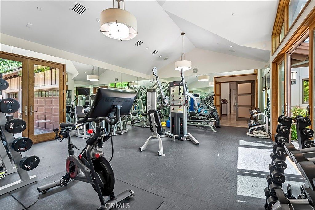This spacious and well-lit home gym features a high vaulted ceiling with modern pendant lighting and large glass doors that provide a view of the outdoors. The room is equipped with a Peloton stationary bike, a multi-station weight machine, and organized racks of UMAX dumbbells, all set against a dark, durable flooring. The expansive mirror wall enhances the sense of space, creating a professional and inviting environment for fitness enthusiasts.
