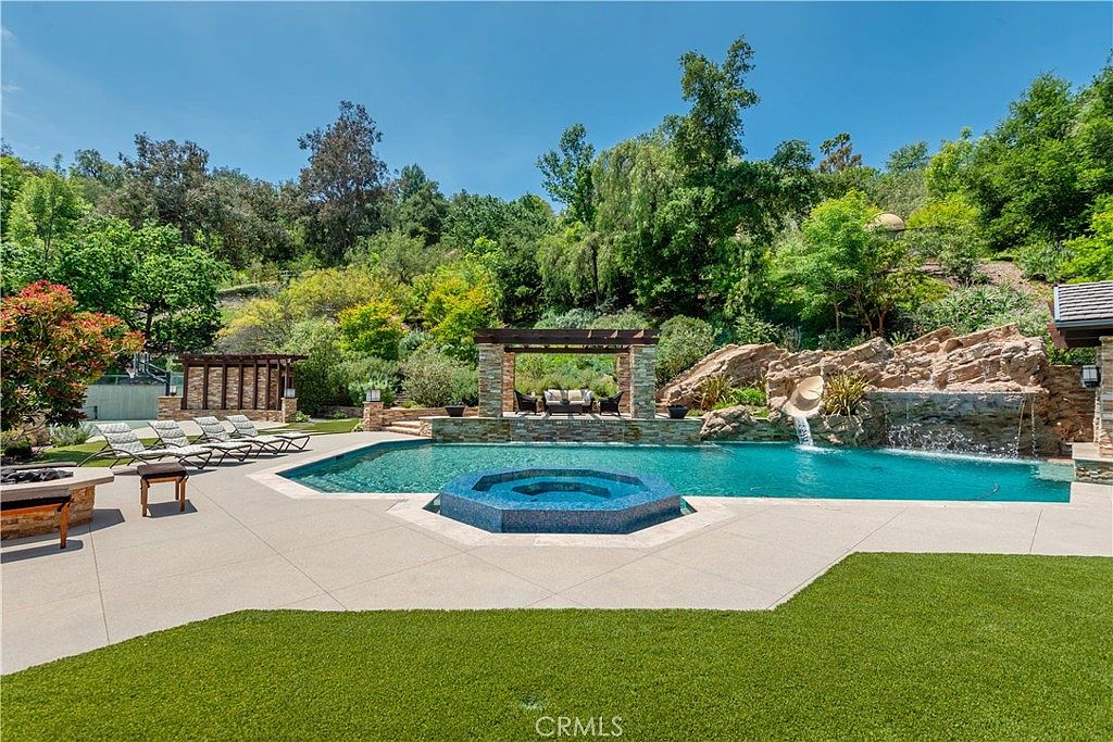 This luxurious backyard oasis features a large, custom-shaped swimming pool with an integrated blue-tiled spa and a natural rock waterfall feature. A covered pergola provides a shaded seating area, while a spacious concrete deck offers ample room for lounge chairs, all set against a lush, landscaped hillside. The scene captures a serene and high-end resort-style atmosphere perfect for outdoor entertaining.