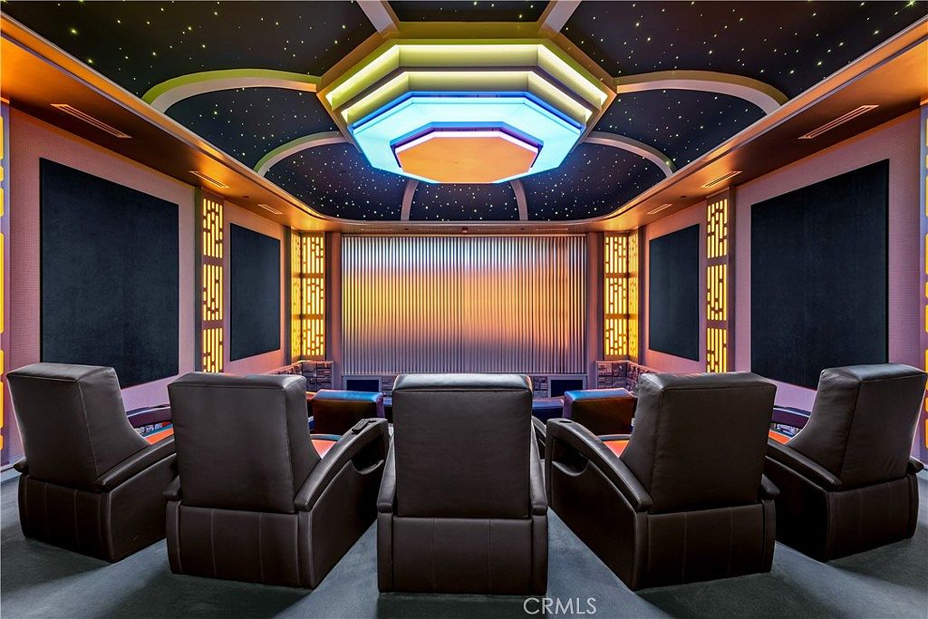 This image showcases a luxurious, custom-designed home theater room featuring five plush, dark brown leather reclining seats arranged in a row. The ceiling is a standout architectural feature, designed to mimic a starry night sky with recessed, multi-colored LED lighting in a tiered, geometric pattern. The room is framed by dark acoustic wall panels and illuminated by vertical, warm-toned light strips, creating an immersive and cinematic atmosphere.