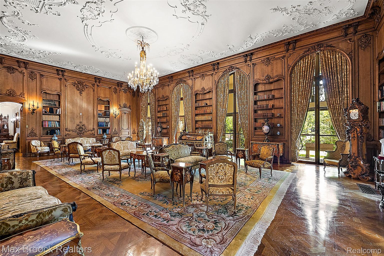 This grand living room features ornate wood paneling, built-in bookshelves, and a stunning chandelier. The room is furnished with elegant, vintage-style seating arranged around a large, decorative rug. Natural light floods the space through large windows draped with luxurious curtains, creating a warm and inviting atmosphere.