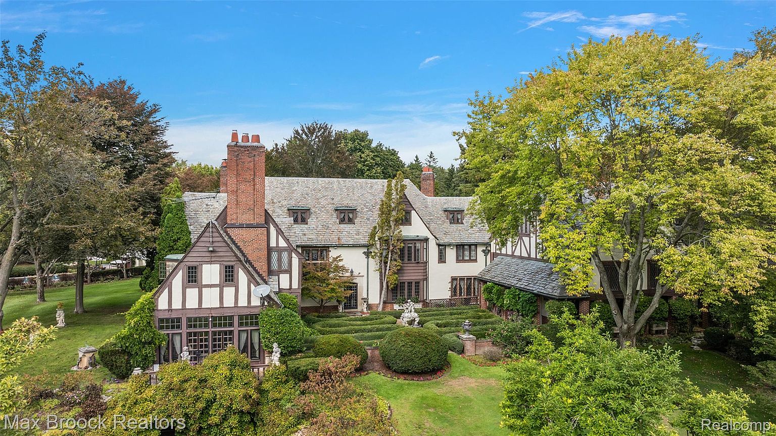 This is an eye-level, cinematic shot of a large Tudor-style home with a meticulously landscaped front yard. The house features a combination of brick and stucco exterior, with prominent gables and multiple chimneys. The surrounding greenery includes mature trees and manicured hedges, creating a sense of privacy and elegance.