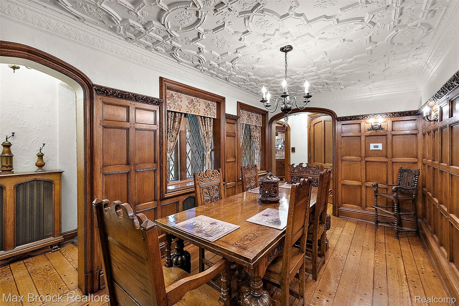This is an interior shot of a formal dining room featuring dark wood paneling, a coffered ceiling, and a large wooden dining table with ornate chairs. The room is illuminated by a chandelier, and natural light filters through the windows. The overall impression is one of classic elegance and traditional style.