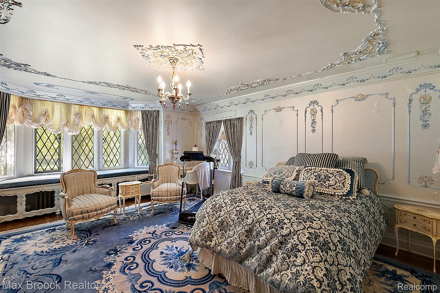 This is a luxurious primary bedroom featuring a large bed with a patterned comforter and decorative pillows. The room is adorned with ornate ceiling and wall moldings, complemented by a chandelier and a patterned rug. A bay window area with seating and two armchairs creates a cozy and elegant atmosphere.