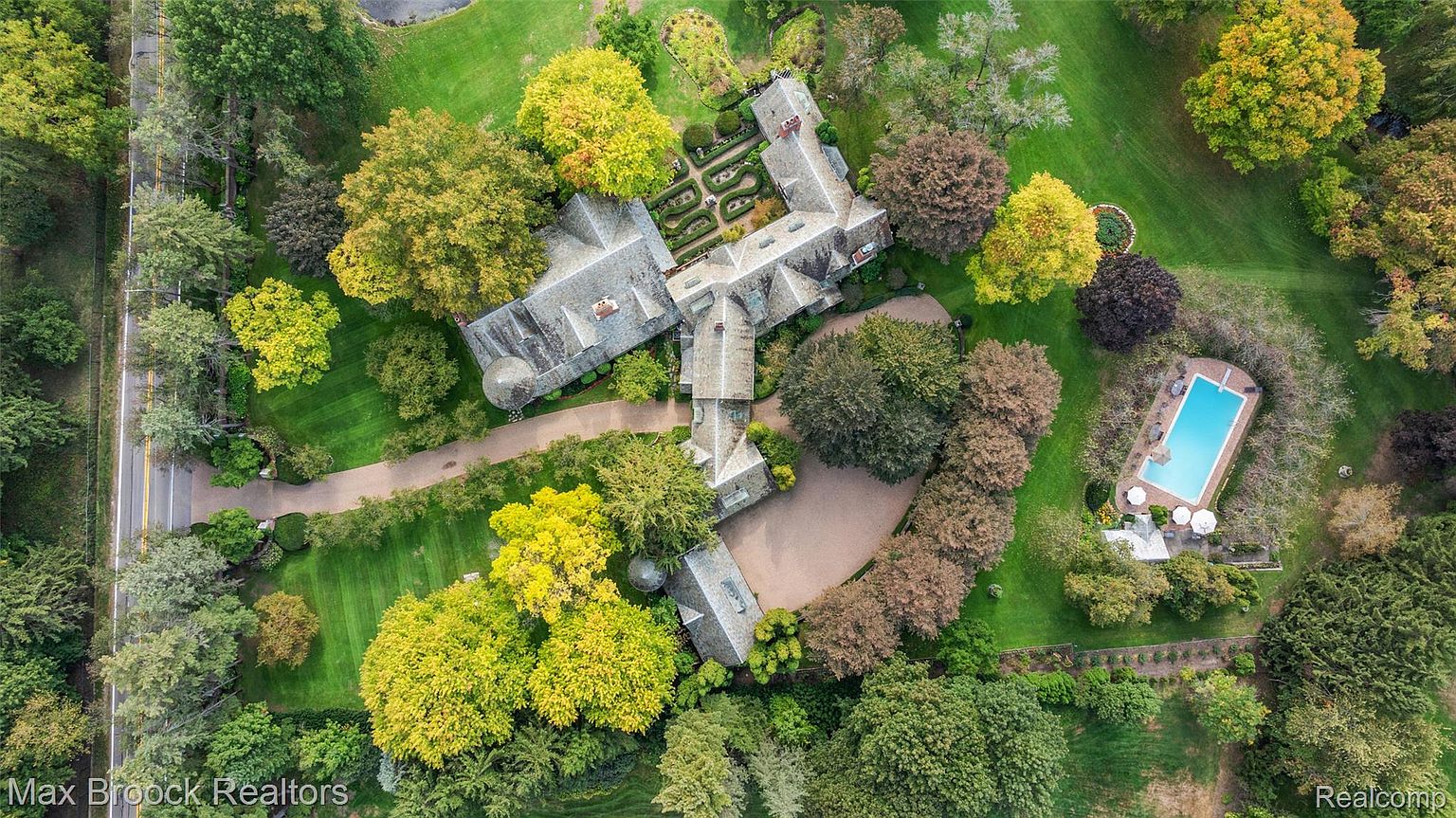 This aerial shot showcases an expansive estate with a sprawling residence, meticulously landscaped gardens, and a private swimming pool. The house features a complex roofline and multiple wings, suggesting a luxurious and well-designed property. Mature trees surround the property, providing privacy and a sense of seclusion.