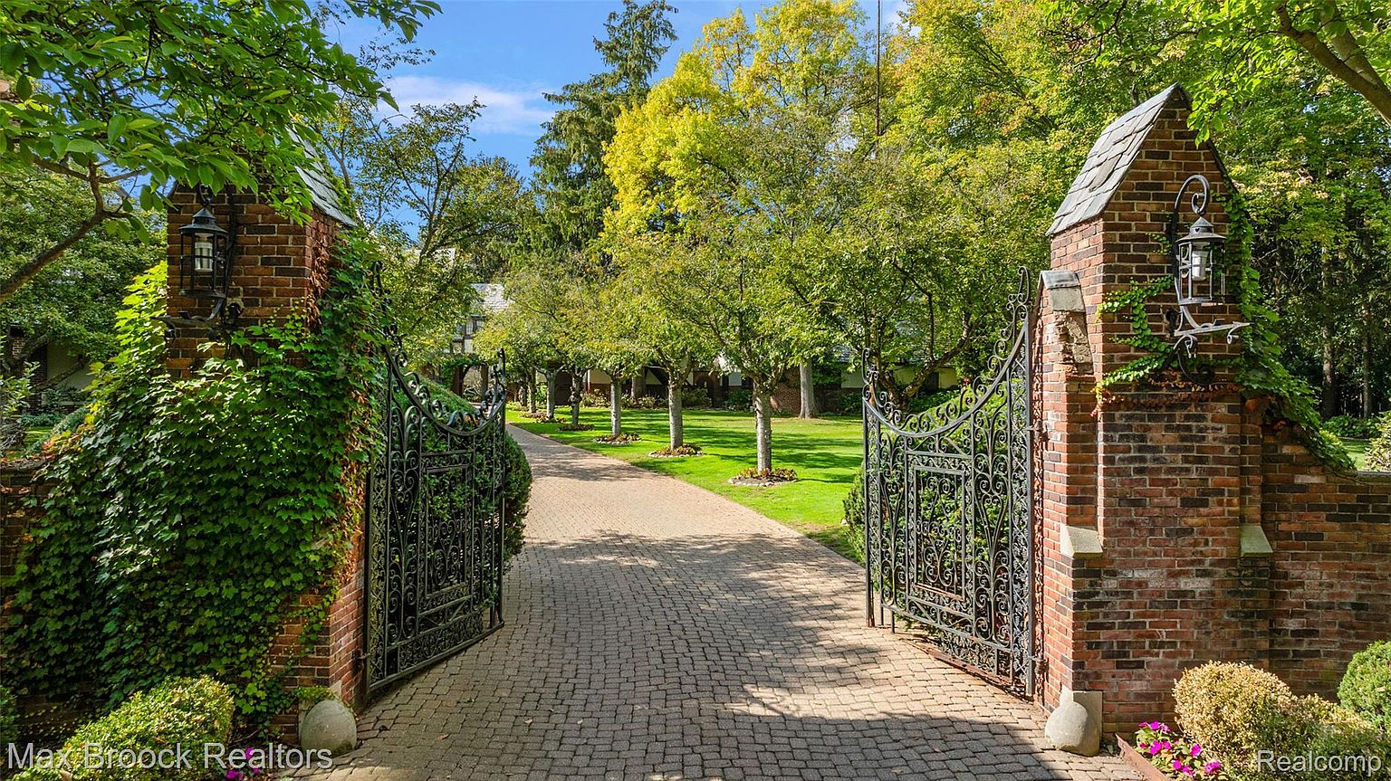 This image showcases a grand entryway featuring ornate wrought iron gates flanked by brick pillars adorned with climbing ivy and vintage-style lanterns. The cobblestone driveway leads through the gates, framed by lush green lawns and mature trees, creating an inviting and impressive approach to the property. The perspective is from the driveway looking into the property.