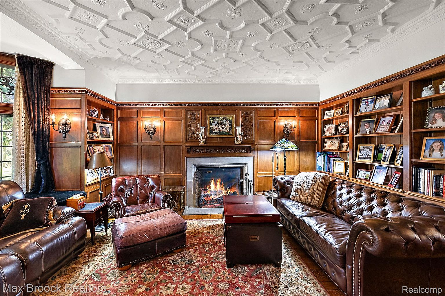 This is a luxurious living room featuring rich wood paneling, built-in bookshelves filled with photos and books, and a coffered ceiling. A leather Chesterfield sofa and armchairs are arranged around a fireplace, creating a warm and inviting atmosphere. The room is illuminated by sconces and lamps, enhancing the classic and sophisticated style.