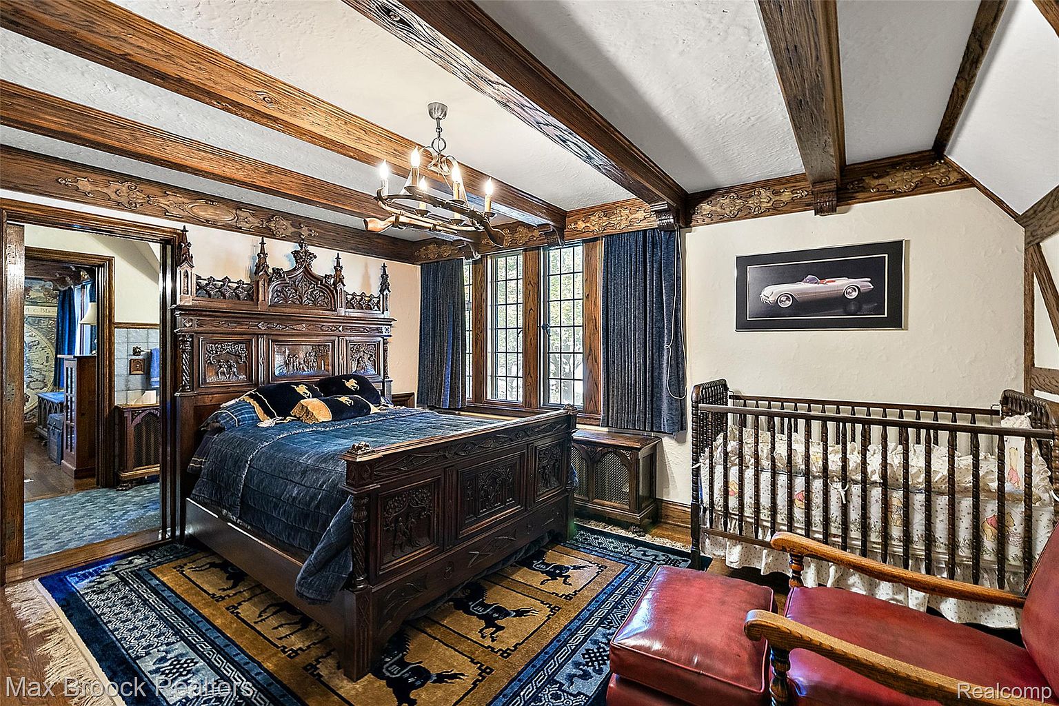 This is a primary bedroom featuring a large, ornate wooden bed with dark blue bedding. The room has exposed wooden beams on the ceiling, a chandelier, and a window with blue curtains. A crib and a framed picture of a car are also visible, adding a personal touch to the space.