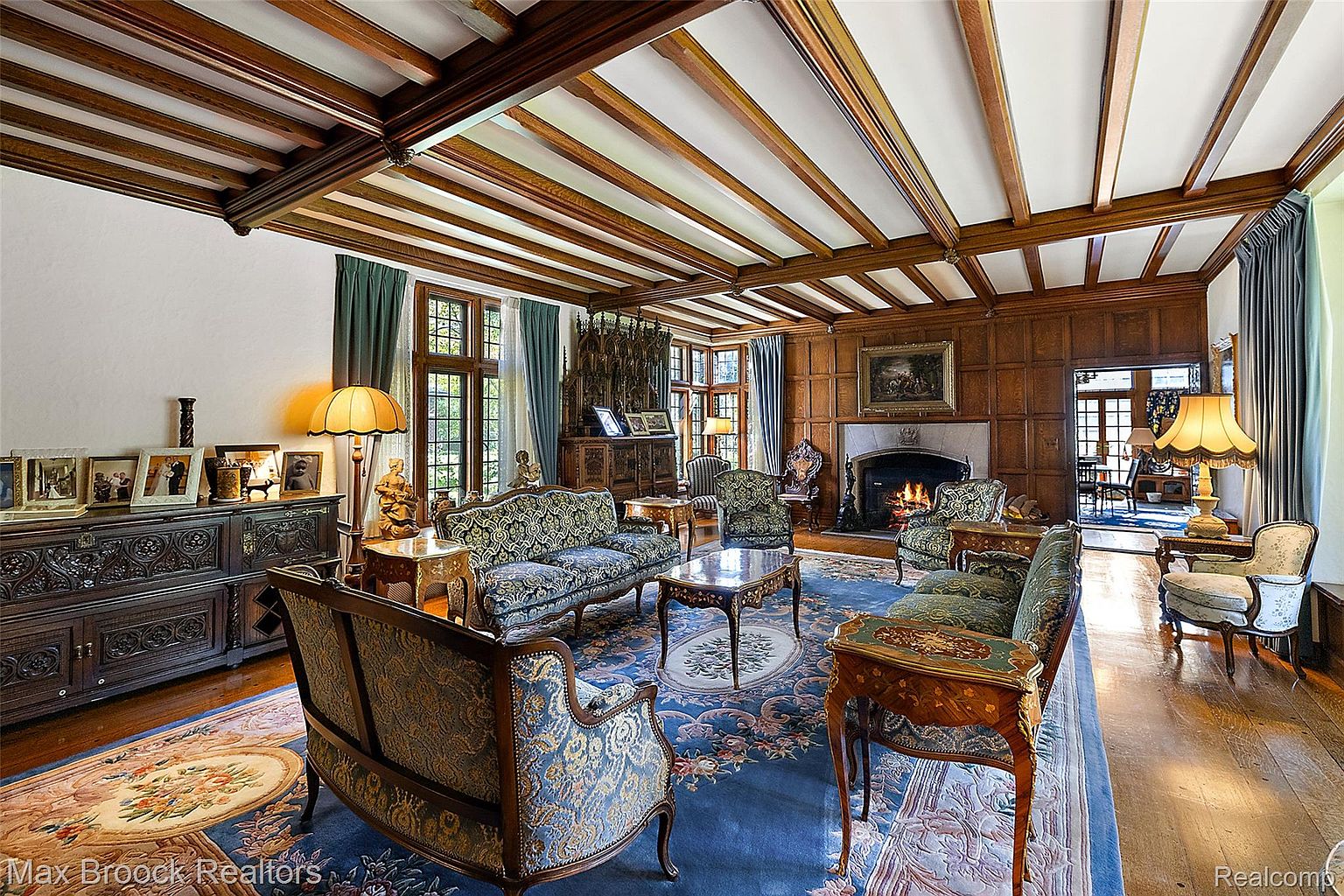 This is a luxurious living room featuring a coffered ceiling with wooden beams, wood-paneled walls, and a fireplace. The room is furnished with ornate sofas and chairs, a patterned rug, and a large wooden cabinet. The overall impression is one of classic elegance and sophistication.