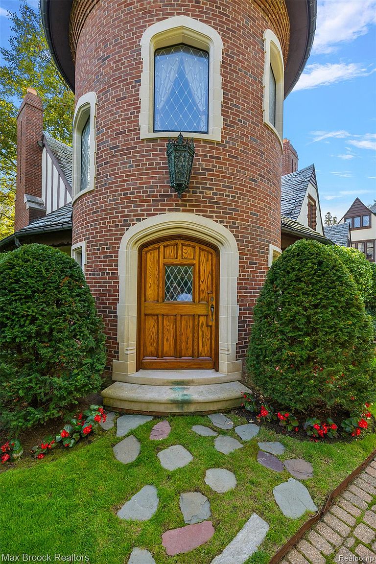 This image showcases the charming entryway of a Tudor-style home, featuring a round brick tower with arched windows and a wooden door with diamond-patterned glass. Lush greenery flanks the entrance, complemented by a stone pathway leading to the door. The overall impression is one of classic elegance and inviting curb appeal.