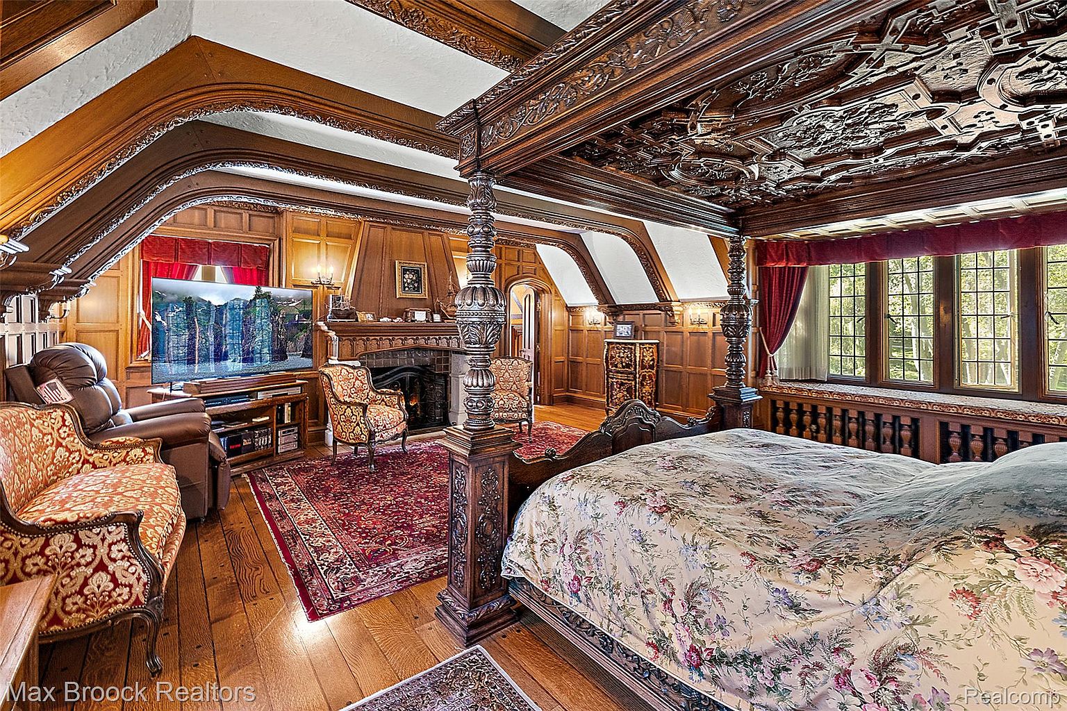 This is a luxurious primary bedroom featuring a four-poster bed with intricate wood carvings and a floral patterned comforter. The room boasts rich wood paneling, a fireplace, and a window seat overlooking the outdoors. The overall impression is one of opulence and old-world charm.