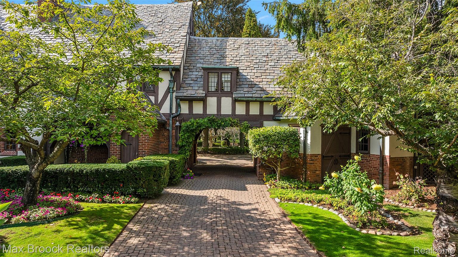 This is a charming front view of a Tudor-style home, showcasing a brick and stucco facade with a slate roof. The property features a brick driveway leading through an arched entryway, lush landscaping with manicured hedges, and mature trees that frame the house. The overall impression is one of classic elegance and well-maintained grounds.