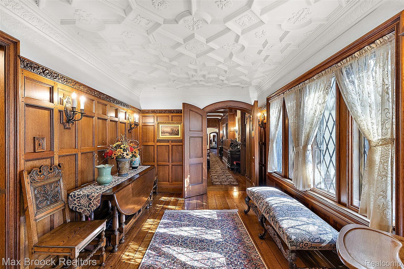 This interior shot showcases a grand hallway featuring rich wood paneling, an ornate coffered ceiling, and hardwood floors. A patterned rug leads the eye down the hallway, while a window seat and antique furniture add to the room's character. The overall impression is one of classic elegance and timeless design.