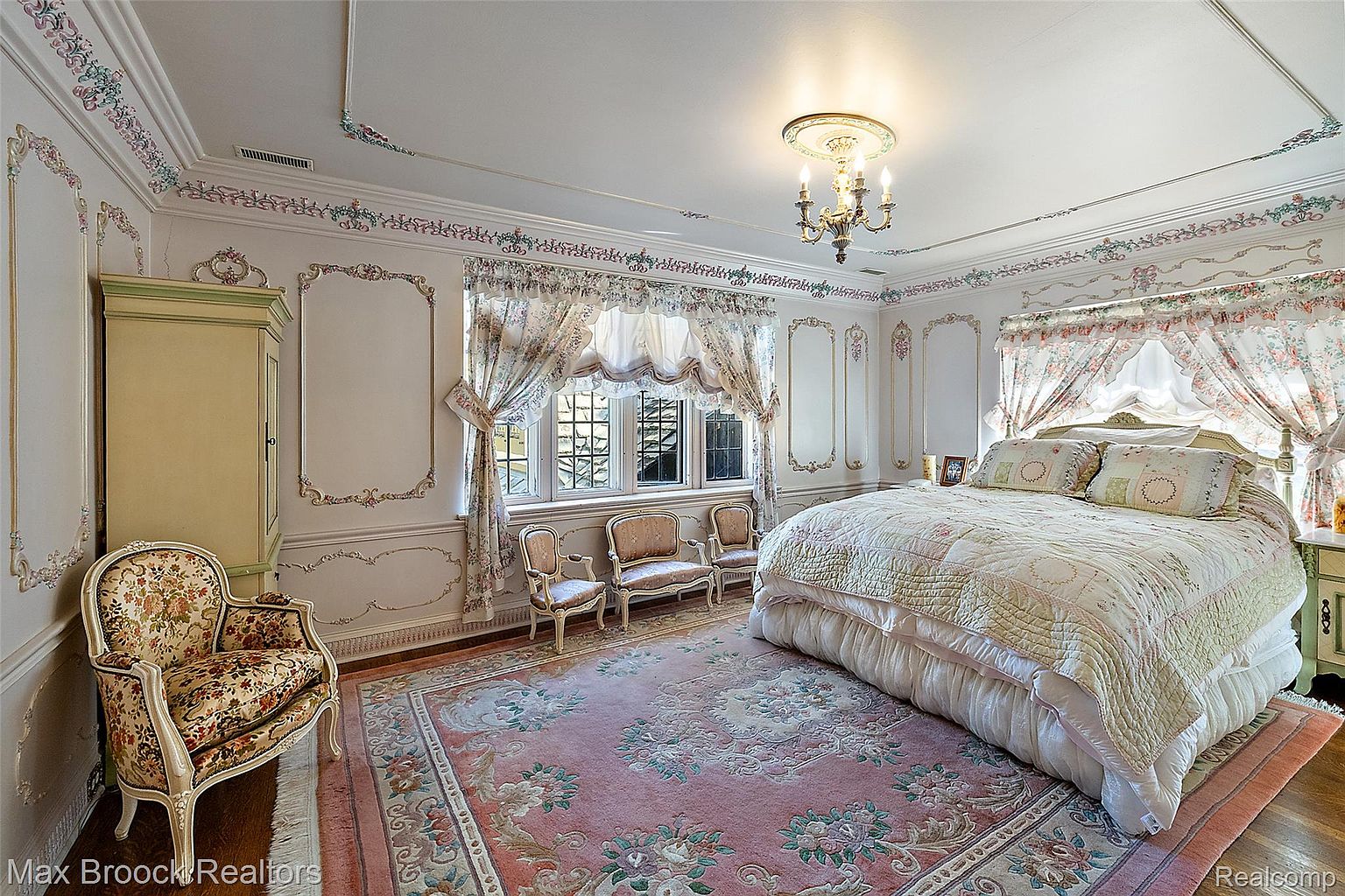 This is a luxurious primary bedroom featuring ornate floral decorations on the walls and ceiling, creating a classic and elegant atmosphere. The room includes a large bed with a floral quilt, a patterned rug, and several antique-style chairs near a window with floral curtains. A vintage chandelier hangs from the ceiling, adding to the room's opulent and traditional style.