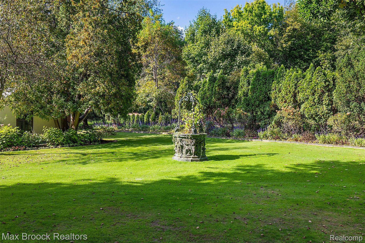 This image showcases a meticulously maintained yard or garden. A central stone fountain, adorned with climbing plants, serves as a focal point, surrounded by a lush green lawn. Mature trees and manicured shrubs frame the space, creating a serene and private outdoor setting.
