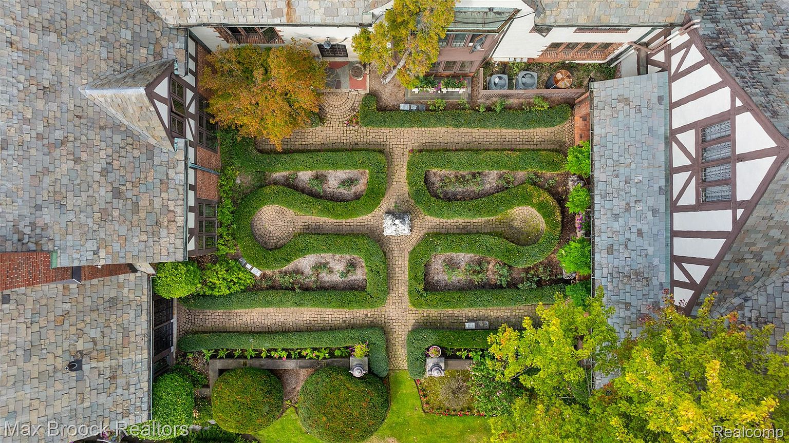 This aerial shot showcases a meticulously designed formal garden nestled within the courtyard of a Tudor-style building. The garden features symmetrical patterns of manicured hedges, brick pathways, and a central sculpture, creating a sense of order and elegance. The surrounding architecture complements the garden's aesthetic, highlighting the property's unique charm and historical character.