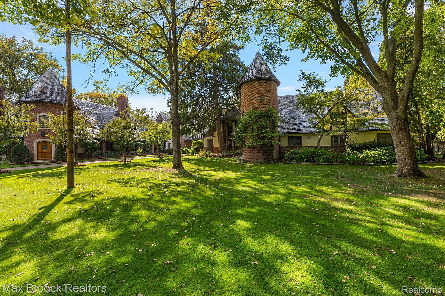 This image showcases a sprawling, well-manicured lawn leading up to a stately home with unique architectural features, including a prominent brick tower and steeply pitched roofs. Mature trees provide ample shade and enhance the property's curb appeal, creating a sense of established elegance and privacy. The overall impression is one of a grand estate with meticulous landscaping.