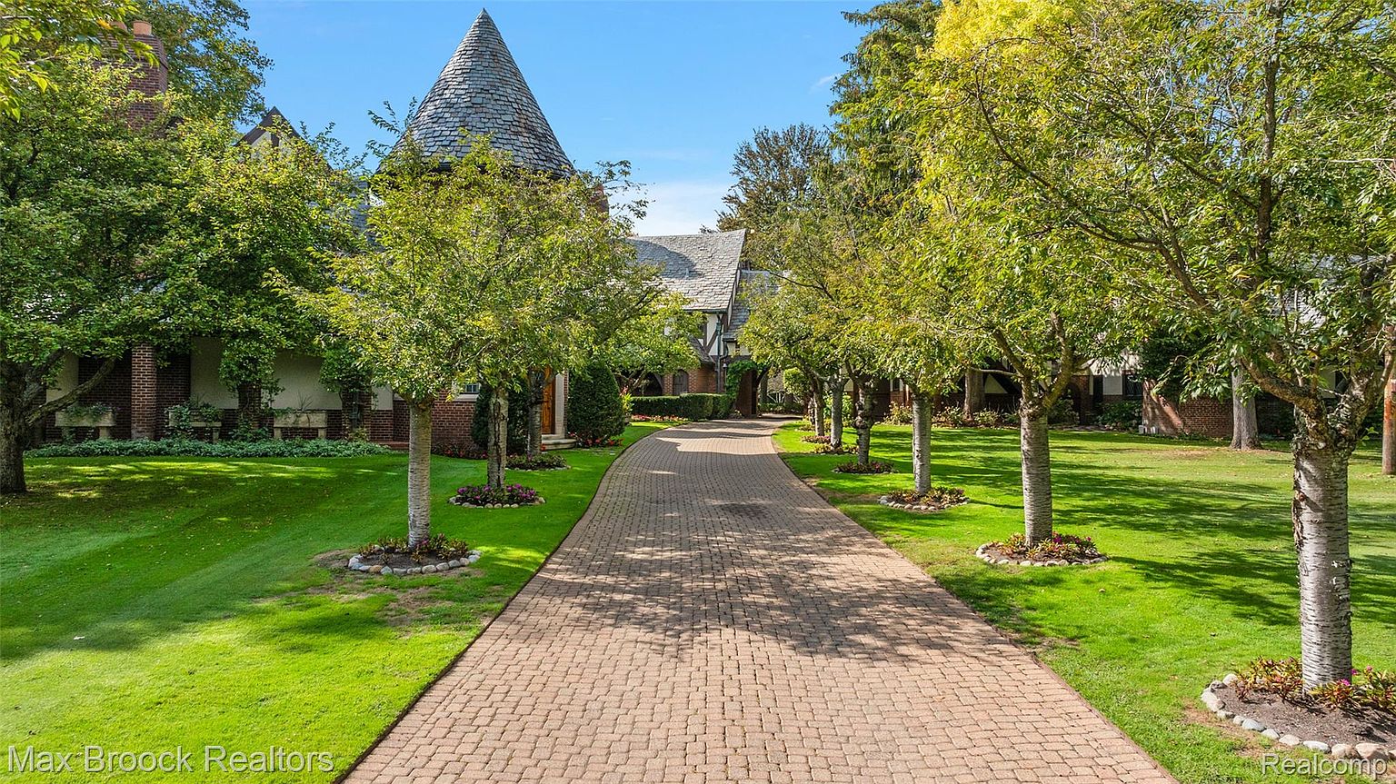 This is a front view of a grand estate, showcasing a long, brick-paved driveway leading to the main entrance. The property features lush green lawns, mature trees lining the driveway, and a unique turret-style architectural element. The overall impression is one of luxury and established elegance.