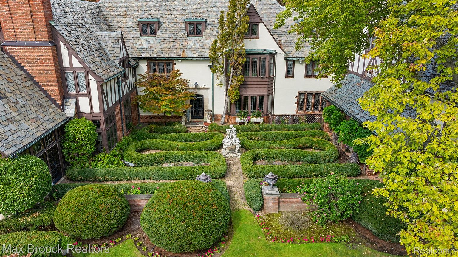 This aerial view showcases a grand Tudor-style home with a meticulously manicured formal garden. The garden features symmetrical hedges, a central statue, and brick pathways, creating a sophisticated and inviting outdoor space. The home's architectural details, including the varied rooflines and window styles, add to its charm and curb appeal.