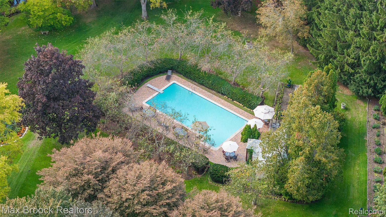 This aerial shot showcases a rectangular in-ground pool surrounded by a brick patio and lush landscaping. The pool area features lounge chairs, umbrellas, and mature trees, creating a private and serene outdoor space. The overall impression is one of luxury and relaxation.