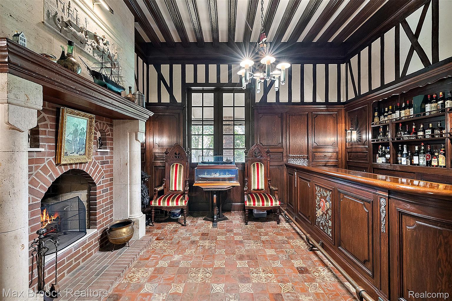 This interior shot showcases a sophisticated wine cellar or bar area, featuring dark wood paneling, a brick fireplace, and a custom-built bar with ample shelving for bottles. Two ornate chairs flank a small table displaying a model ship, adding a nautical touch to the room's classic and elegant design. The room is lit by a chandelier and wall sconces, creating a warm and inviting atmosphere.