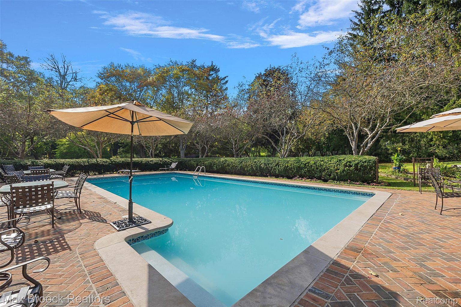 This image showcases a rectangular swimming pool surrounded by a brick patio. A large umbrella provides shade over a seating area with wrought iron furniture. The pool is bordered by a neatly trimmed hedge and mature trees, creating a private and inviting outdoor space.