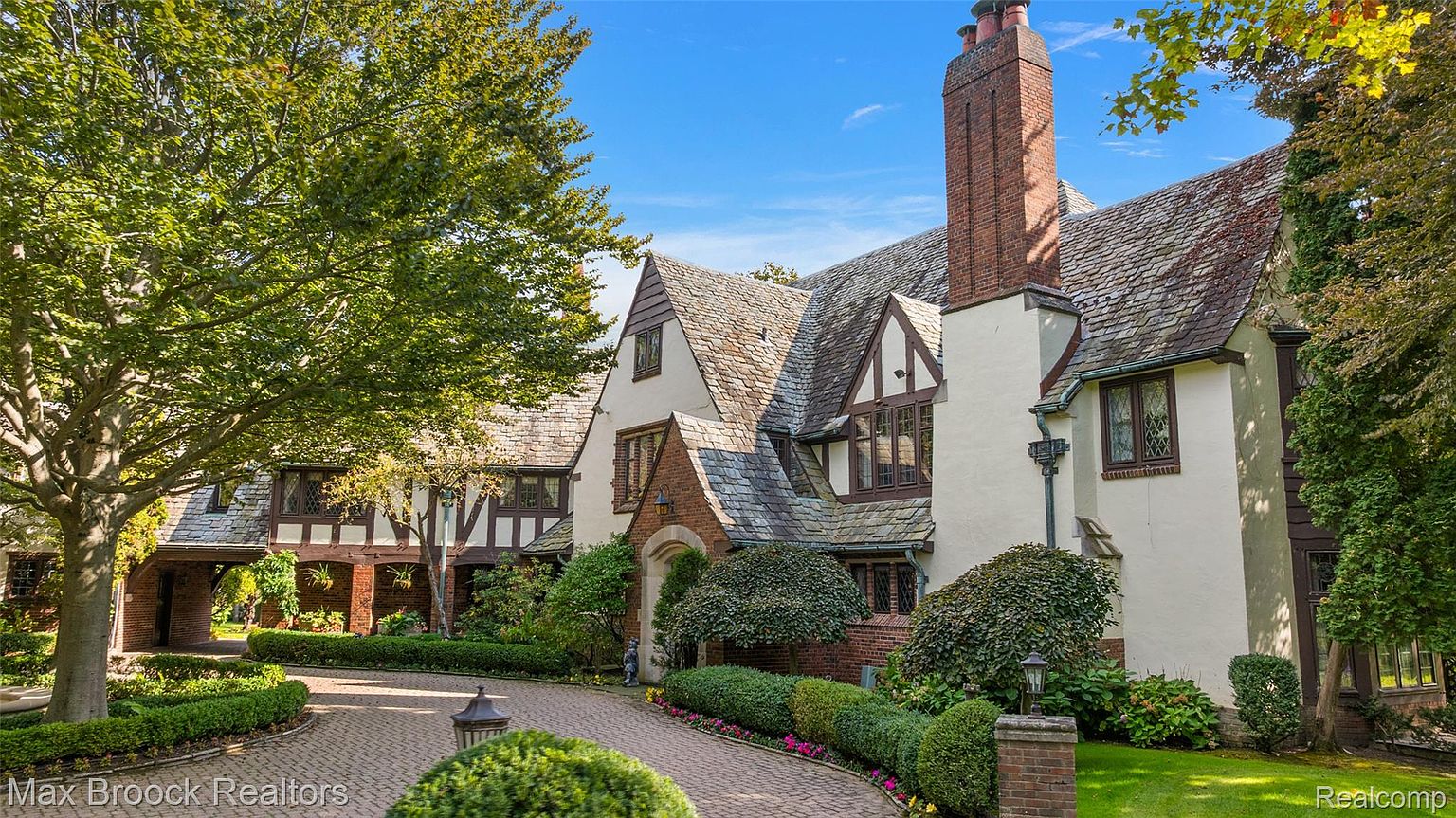 This is a front exterior view of a large Tudor-style home with a brick driveway and meticulously maintained landscaping. The house features a combination of white stucco and dark wood trim, a steeply pitched slate roof, and a prominent brick chimney. The overall impression is one of classic elegance and established luxury.