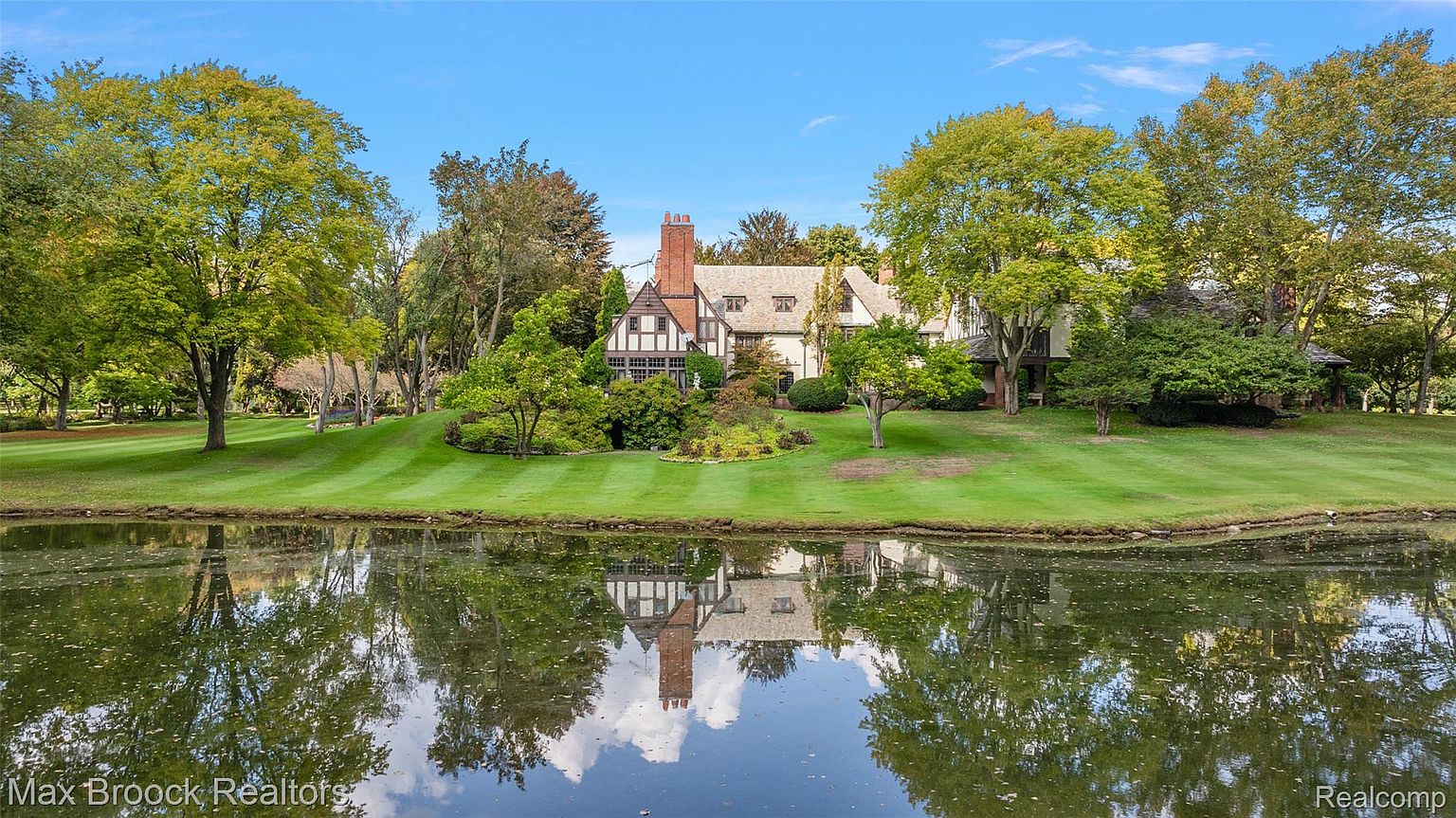 This image showcases the front exterior of a grand Tudor-style home, reflected beautifully in a serene pond. The meticulously manicured lawn and mature trees frame the house, creating a picturesque and inviting scene. The architectural details, including the brick chimney and gabled roof, add to the home's charm and curb appeal.