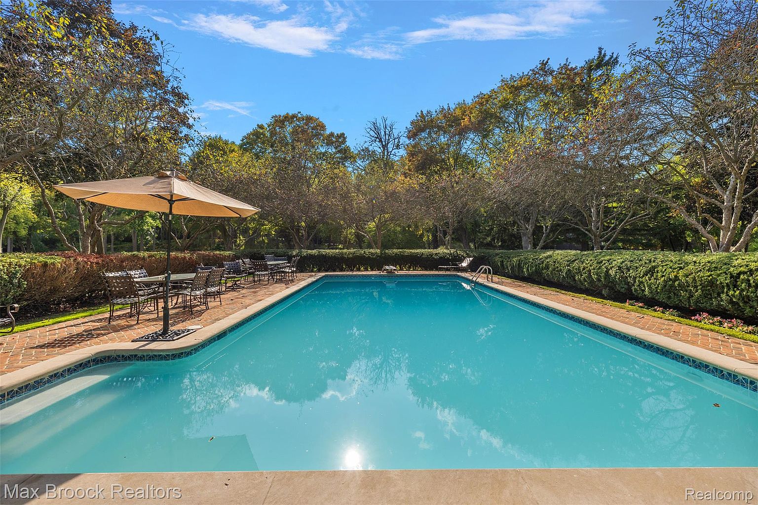 This image showcases a pristine swimming pool surrounded by lush greenery and mature trees, creating a serene and private backyard oasis. The pool features a tiled edge and clear blue water, reflecting the sky above. An umbrella-covered dining area sits nearby, suggesting a perfect spot for relaxation and outdoor entertaining.