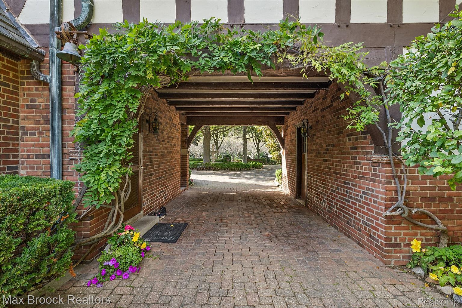 This image showcases a charming brick-paved entryway leading into a garden area. The entrance is framed by brick walls adorned with lush greenery, creating a welcoming and picturesque scene. The overhead structure features wooden beams, adding a rustic touch to the overall design.