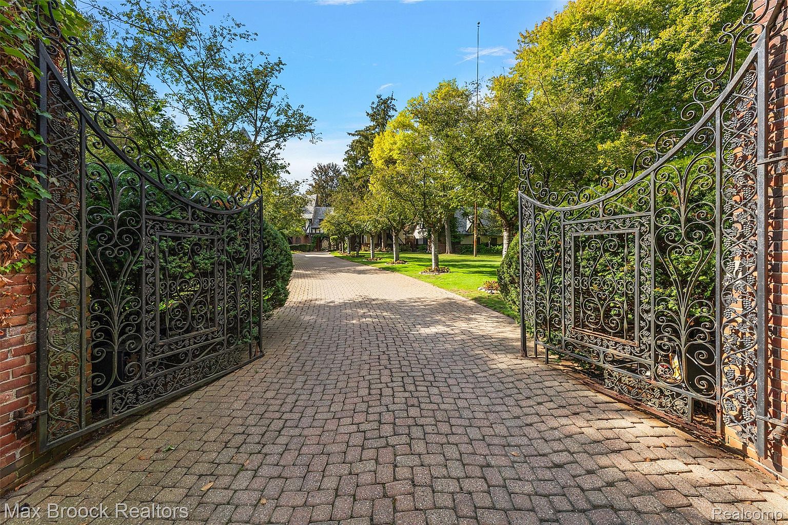 The image showcases a grand entryway to a property, featuring ornate wrought iron gates that are wide open, inviting entry. A brick-paved driveway leads through the gates, lined with mature trees and manicured lawns. The perspective is from the entrance, emphasizing the depth and grandeur of the estate.
