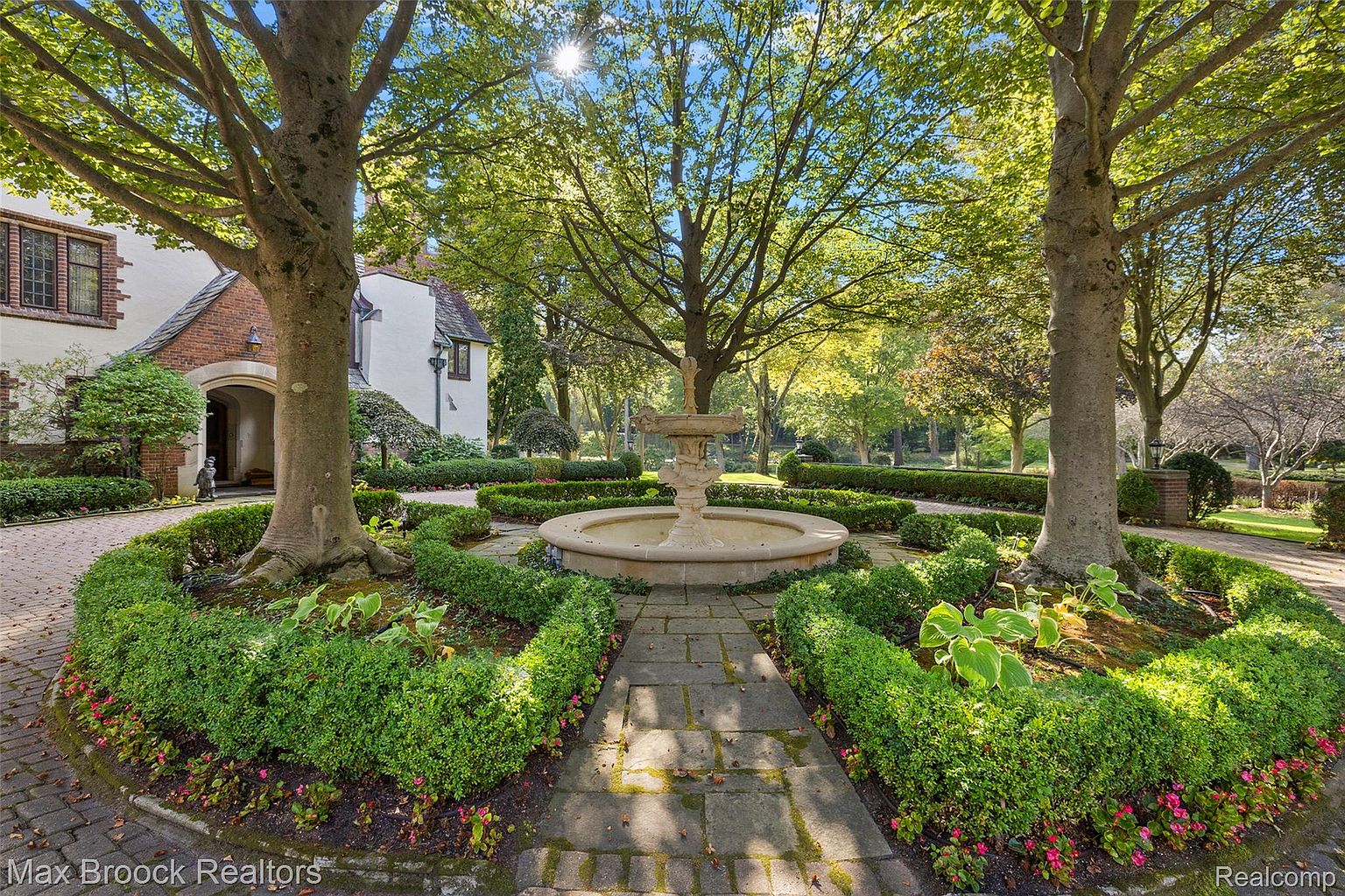 This image showcases a meticulously landscaped garden featuring a central fountain, surrounded by manicured hedges and mature trees. A stone pathway leads to the fountain, enhancing the garden's symmetrical design. The overall impression is one of tranquility and sophisticated outdoor living, perfect for a high-end property.