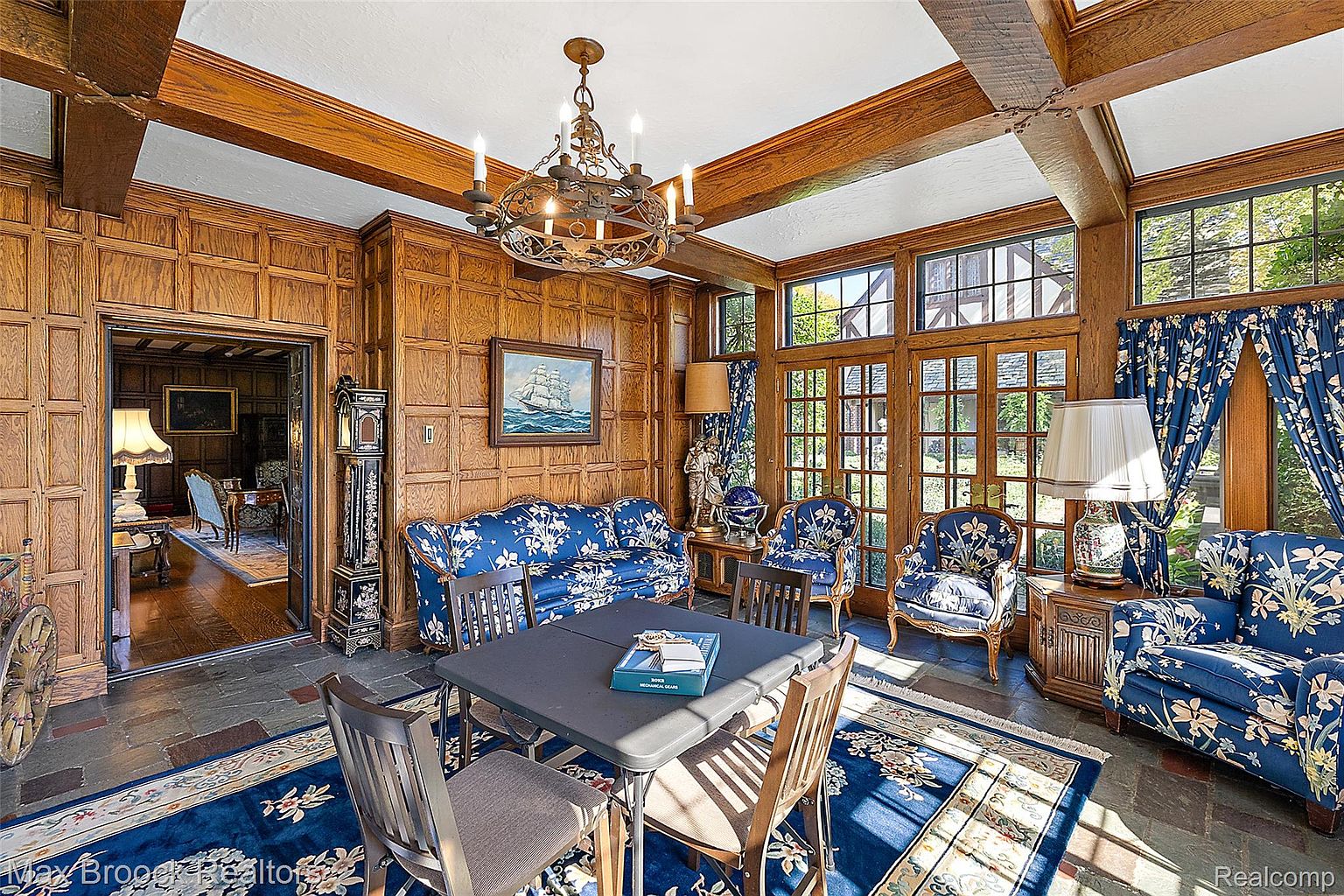 This is an interior shot of a sunroom or conservatory, featuring rich wood paneling and exposed beams. The room is furnished with blue floral patterned sofas and chairs, arranged around a dark folding table. Large windows provide ample natural light, and a blue floral rug complements the furniture, creating a cozy and elegant atmosphere.