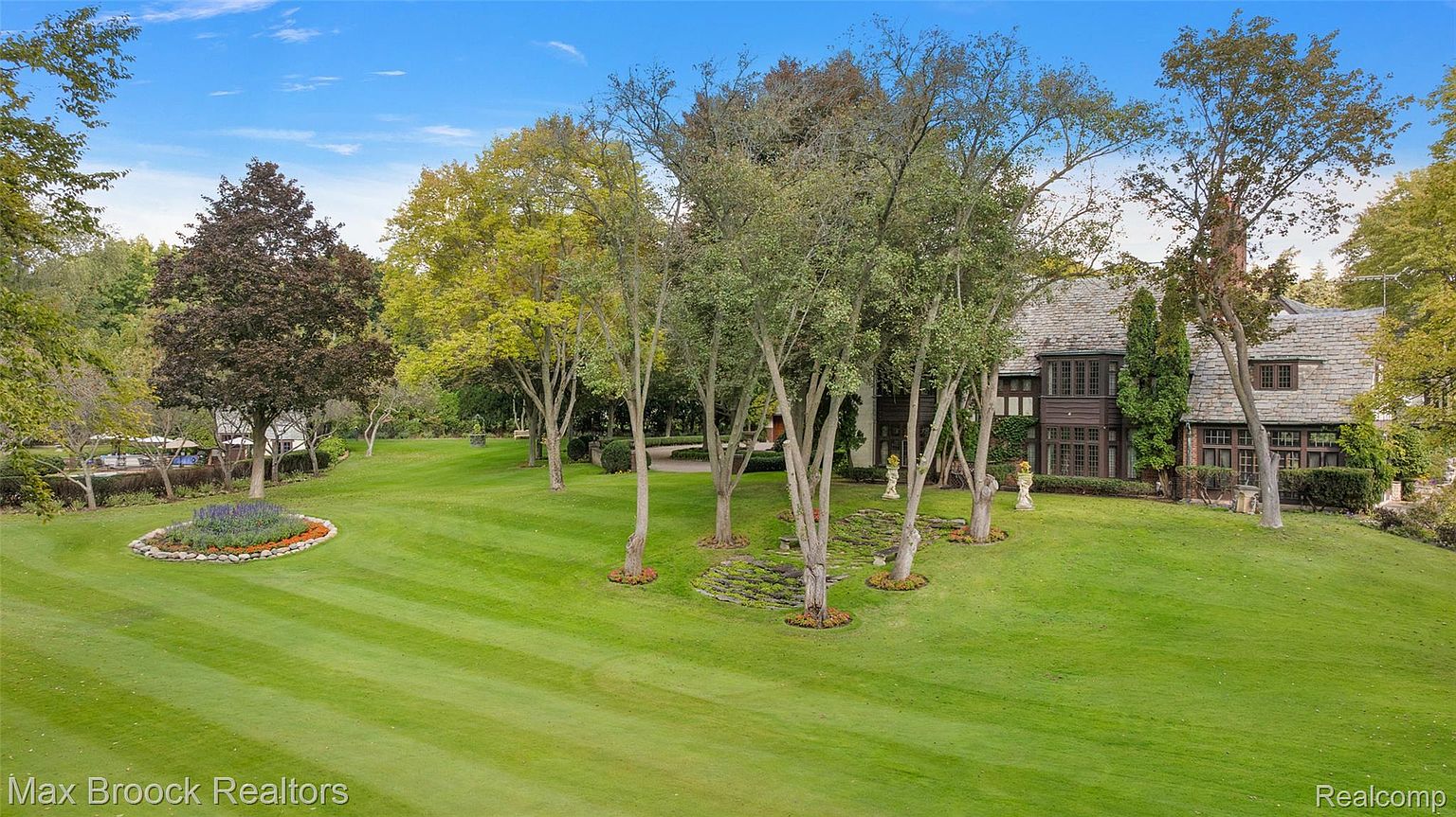 This image showcases a meticulously maintained yard and garden, featuring lush green lawns with striped mowing patterns, mature trees providing shade and visual interest, and a flower bed adding a pop of color. The property includes a large house with a stone roof and dark wood trim, partially obscured by the trees, suggesting a private and luxurious setting. The perspective is a wide shot, emphasizing the expansive and well-manicured landscape.