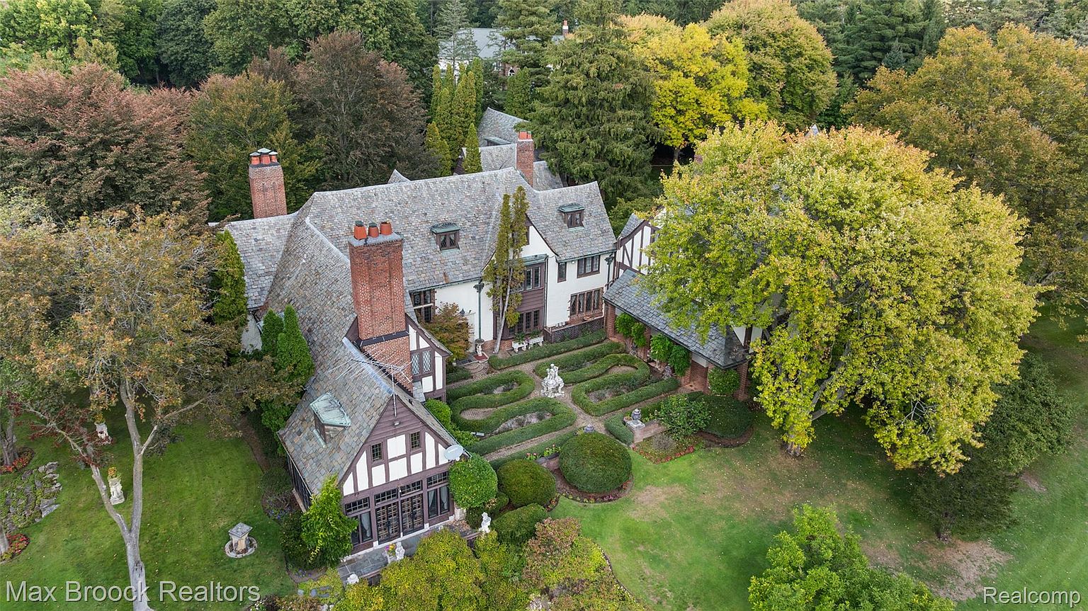 This aerial view showcases a grand Tudor-style home nestled among mature trees and manicured gardens. The property features a complex roofline with multiple gables, brick chimneys, and a formal garden with sculpted hedges and statues. The overall impression is one of established elegance and privacy.