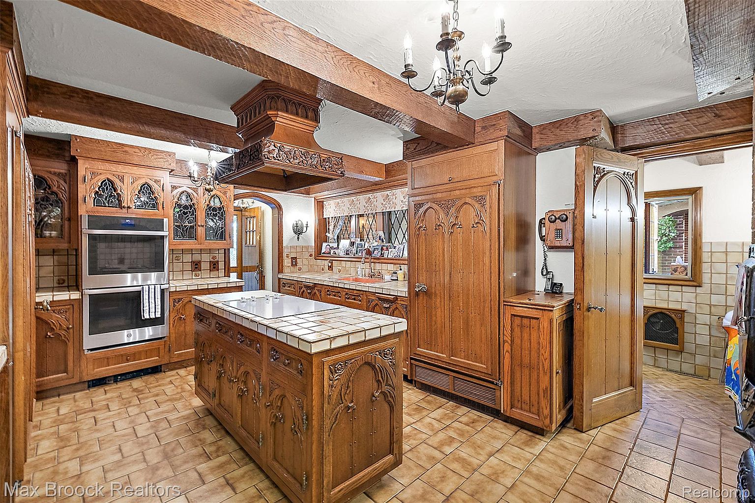 This is a kitchen with a unique, old-world style, featuring custom wood cabinetry with intricate carvings and leaded glass accents. The kitchen island has a tiled countertop, and the room is illuminated by a chandelier and recessed lighting. The overall impression is one of craftsmanship and character.