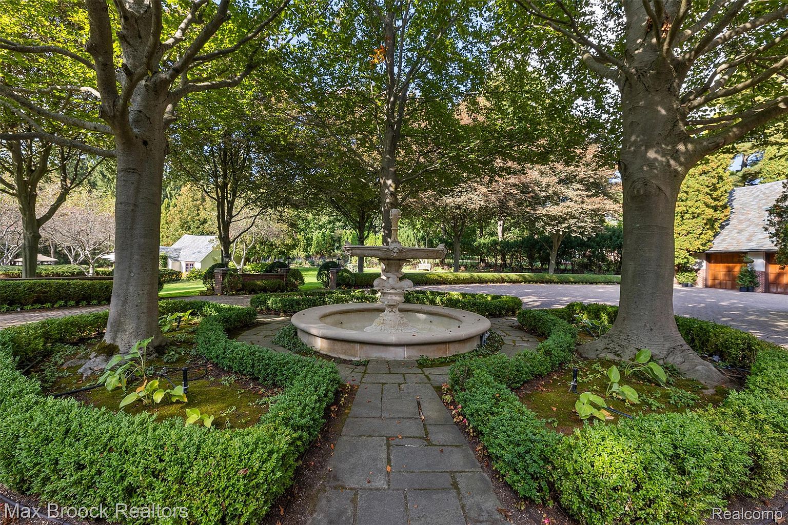 This image showcases a beautifully landscaped garden featuring a circular stone fountain as its centerpiece. The fountain is surrounded by meticulously maintained boxwood hedges and mature trees, creating a serene and elegant outdoor space. A stone pathway leads to the fountain, enhancing the garden's inviting and symmetrical design.